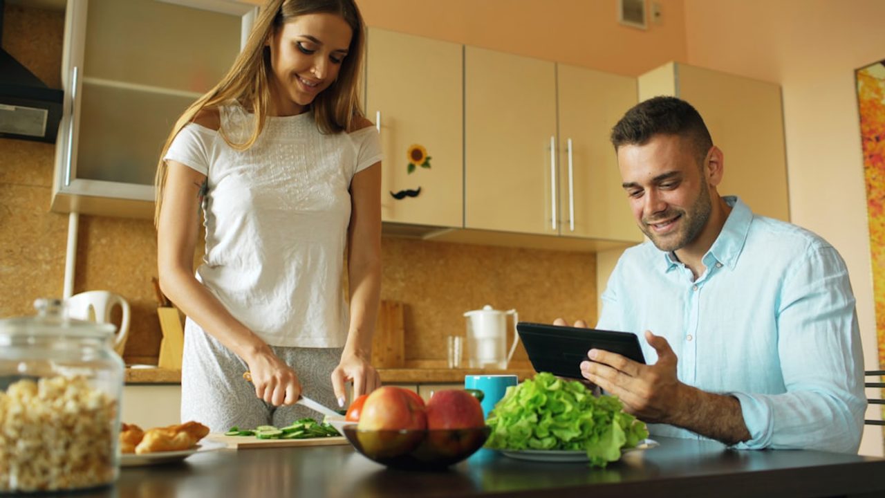 Çekici çift sabahın erken saatlerinde mutfakta sohbet ediyor. Yakışıklı adam kız arkadaşı evde kahvaltı hazırlarken tablet kullanıyor