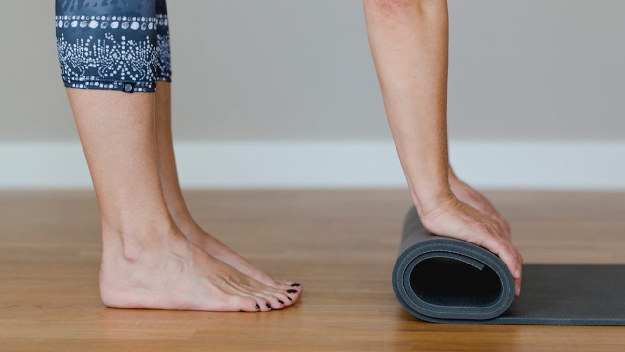 A woman is rolling up her yoga mat in a yoga studio. While doing this, she stands upright and keeps her feet firmly planted on the ground. The yoga mat is blue, and it’s the end of that day’s yoga class.