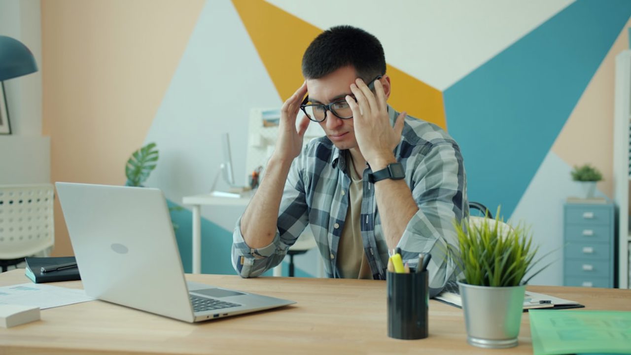 A stressed man with glasses suffers from an overwork-related headache while working on a laptop in the office; he touches his head and rubs his temples. Work and health concept.