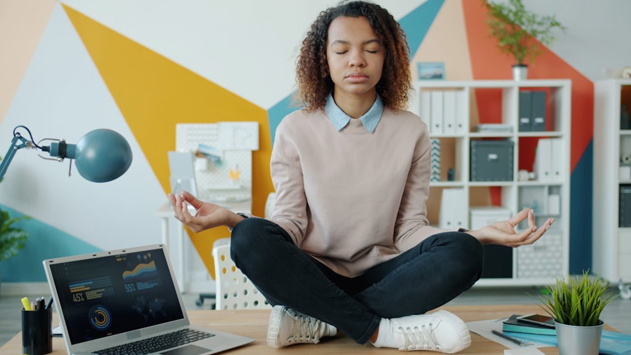 A young African American girl sits in lotus position at an office desk, enjoying relaxation and meditation. Work, stress management, and taking a break at work concept.