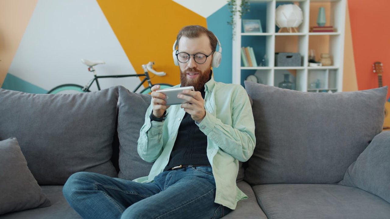 Portrait of a happy young man playing a mobile video game on a smartphone while relaxing alone on a couch at home. Youth and lifestyle concept.