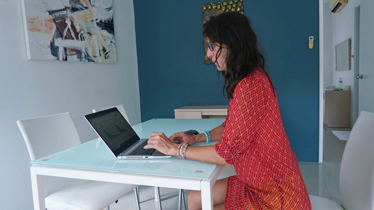 A freelance businesswoman in a red dress works from home, using web-based and mobile technologies to produce instantly updatable and shareable content for training programs. The design of her apartment is minimal and comfortable, helping her focus on tasks.
