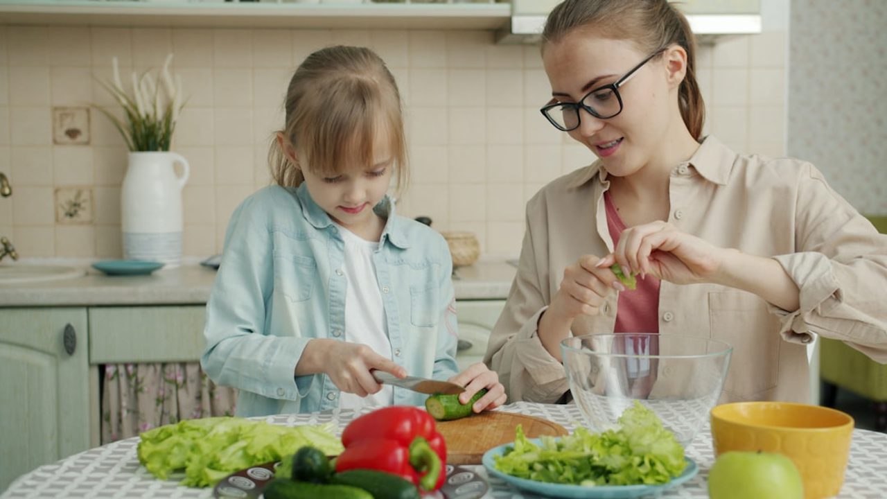 A cute little girl is helping her mother in the kitchen by cutting vegetables for a salad and chatting as they prepare dinner together. Healthy eating, lifestyle, and people concept.
