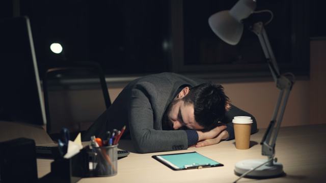 A tired man in a formal suit sleeping on his desk in the office late at night after an intense workday. An open laptop, packaged coffee, and a desk lamp are visible on the desk.
