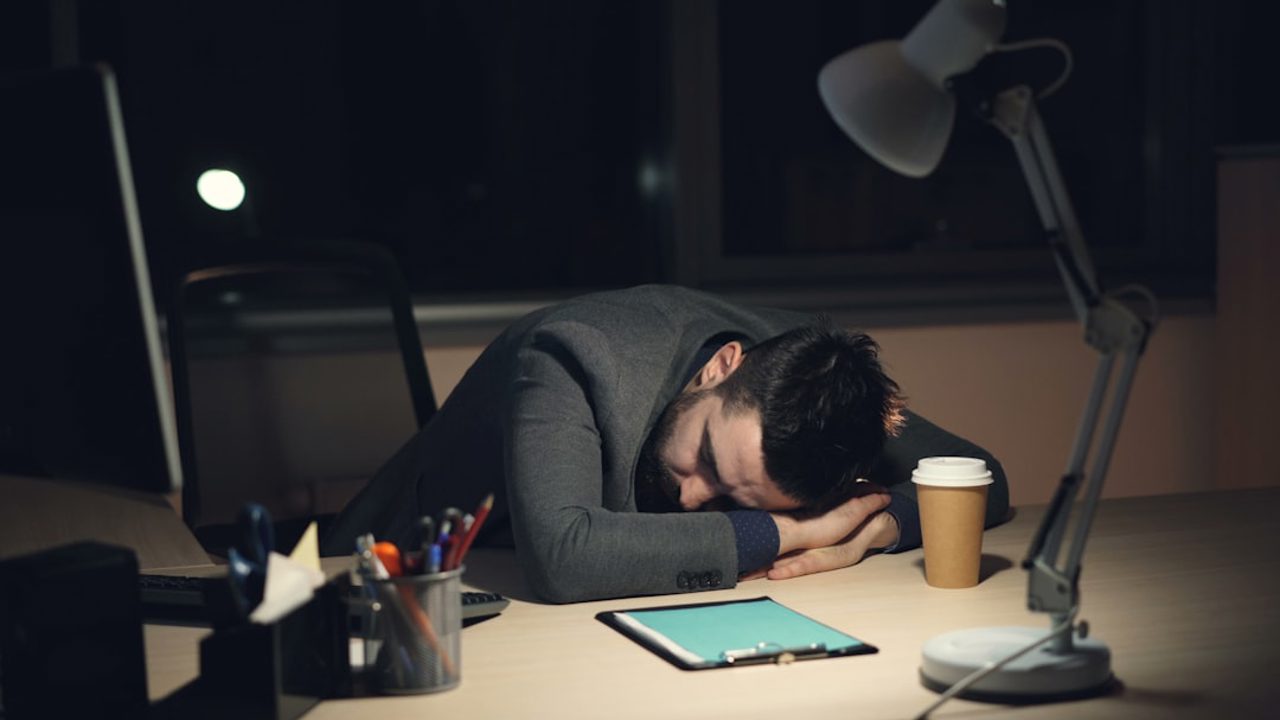A tired man in a formal suit sleeping on his desk in the office late at night after an intense workday. An open laptop, packaged coffee, and a desk lamp are visible on the desk.