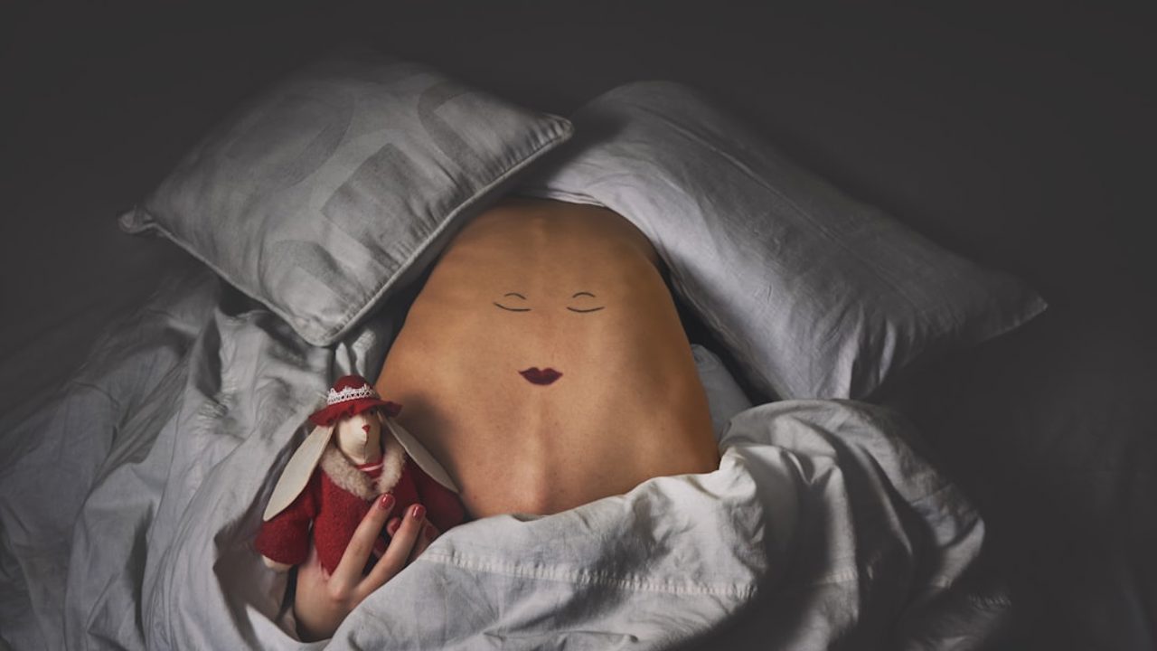 Eine Person liegt unter einer weißen Bettdecke, auf dem Bauch ist ein schlafendes Gesicht gemalt und in der Hand hält sie ein rot gekleidetes Stoffkaninchen.