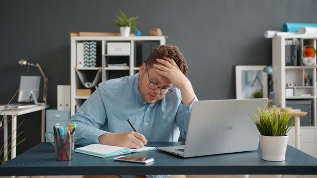 The tired young man is typing and working at a computer indoors at the workplace; he is experiencing a headache, touching his face and head. The concept of workaholism, work, and human health.